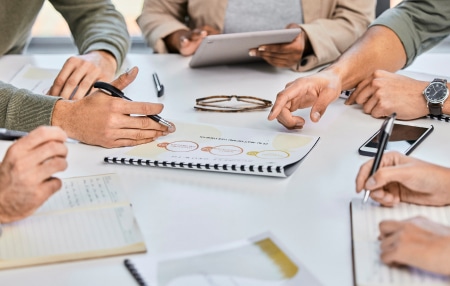 Tips to tackle your debt in 2024 feature view of the hands of five people sitting around a table with a notebook with figures on in the centre of the table