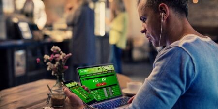 man sat at laptop with mobile in hand. Both device screens display a gambling app