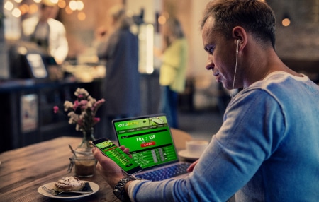 Are-gambling-debts-enforceable_-Feature man sat at laptop with mobile in hand. Both device screens display a gambling app