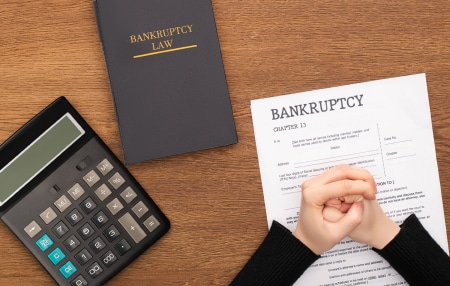 How long does Bankruptcy last in the uk feature birds eye view of brown wooden table with calculator, black book, and bankruptcy document with persons clasped hands placed on top