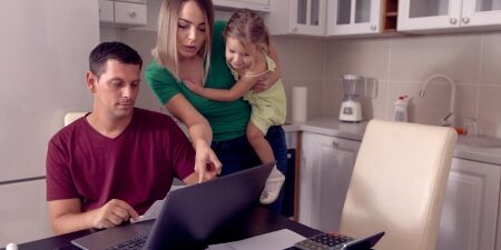 Budget Basics Every Parent Should Know feature An image of a man and a woman holding a baby in their kitchen, pointing at the laptop looking at their budget.