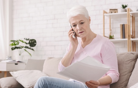 How-to-negotiate-your-bills-feature Middle-aged women sitting on her sofa with documents in one hand and speaking on the phone with the other.