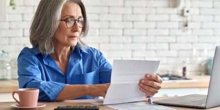 Can-You-Retire-if-You-are-In-Debt-feature An image of a lady who could be at retirement age, sitting at a desk with a coffee, laptop, and reading forms.