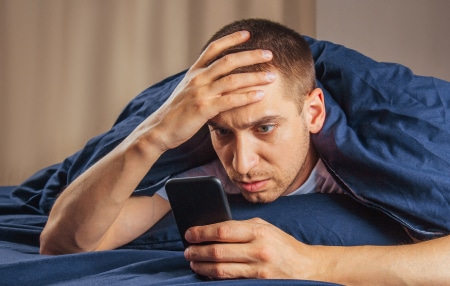 Does-Social-Media-Put-Your-Finances-Under-Pressure_-feature Image of a man lying down in bed on his mobile, looking concerned