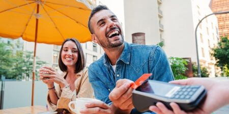 Image of a man and a woman smiling sitting outside of a restaurant paying for their coffee