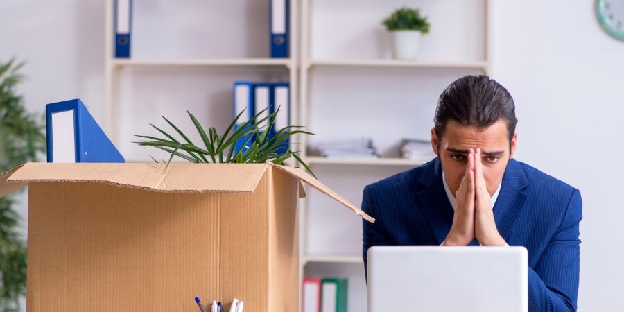 How-To-Cope-With-Job-Loss_-Dealing-With-Debt-After-Redundancy-feature-1 An image of a man sat at his desk with his head in his hands.