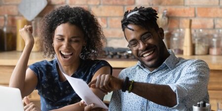 Image of a couple sitting at their table with their laptop, smiling at sheets of paper.