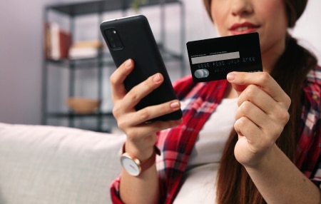 4 Practical Tips for Avoiding Impulse Purchases feature Image of a woman sitting on her sofa with her credit card in one hand and her phone in the other.