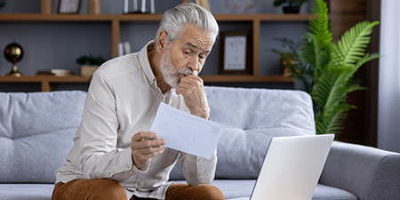 Image of an older man sitting on his sofa reading a letter with his laptop.