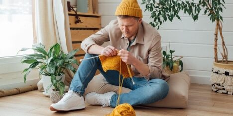 A man in a yellow beanie knitting on the floor in a cozy room.