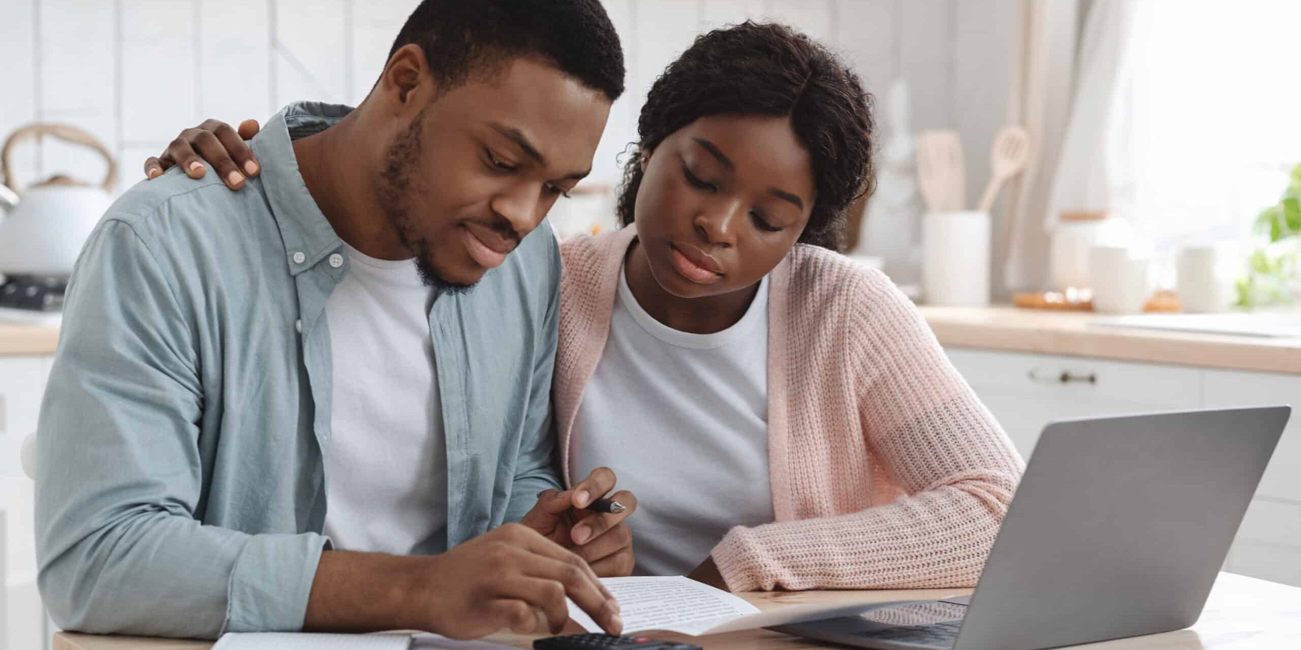 Portraif Of Young African Couple Sitting At Table In Kitchen, Reading Documents A couple reviews documents together at a kitchen table with a laptop and calculator.
