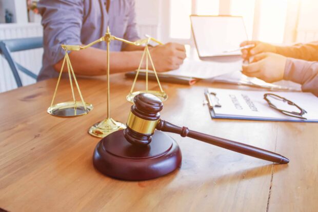 A wooden gavel and golden scales on a desk, with two people reviewing legal documents in the background.