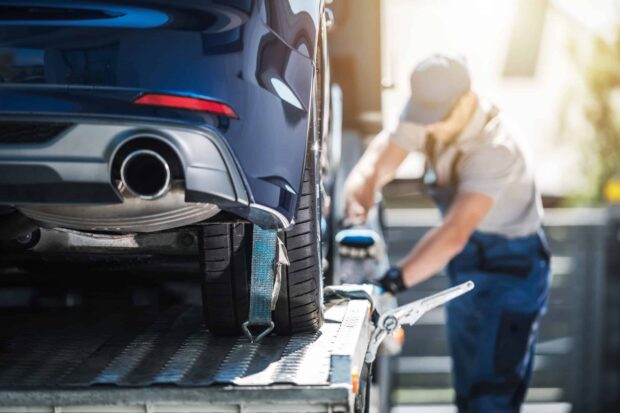 A blue car being secured onto a flatbed tow truck as a worker tightens the strap on the rear tire.