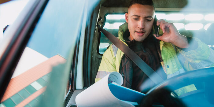 Mid-adult manual worker is sitting in his van, looking through paperwork while speaking to someone on the phone.
